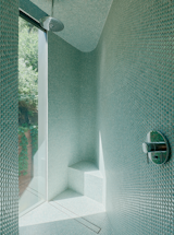 The wedge-shaped shower in the primary bathroom glimmers with sunlight from a full-height window. The tiles are from SomerTile, and the fixtures are from Grohe.