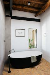 The bathroom blends sleek contemporary fixtures with natural materials. A free-standing black and white tub is surrounded by concrete walls, terra-cotta tile flooring, and an exposed wood ceiling.